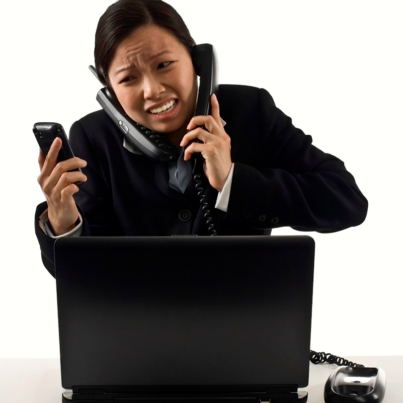 lady stressed holding a cell phone, two land line phones pressed up to her ears, and standing in front of a laptop.